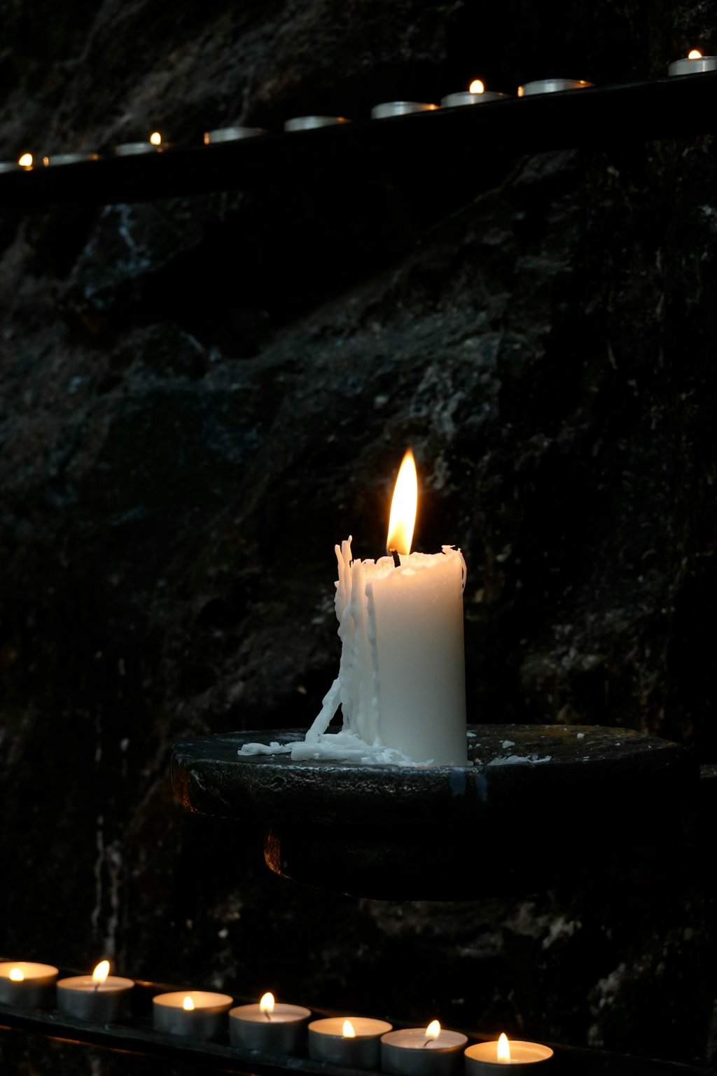 A white pillar candle burning with melted wax drips, surrounded by small tealight candles against a dark stone background. A symbol of remembrance and peace.