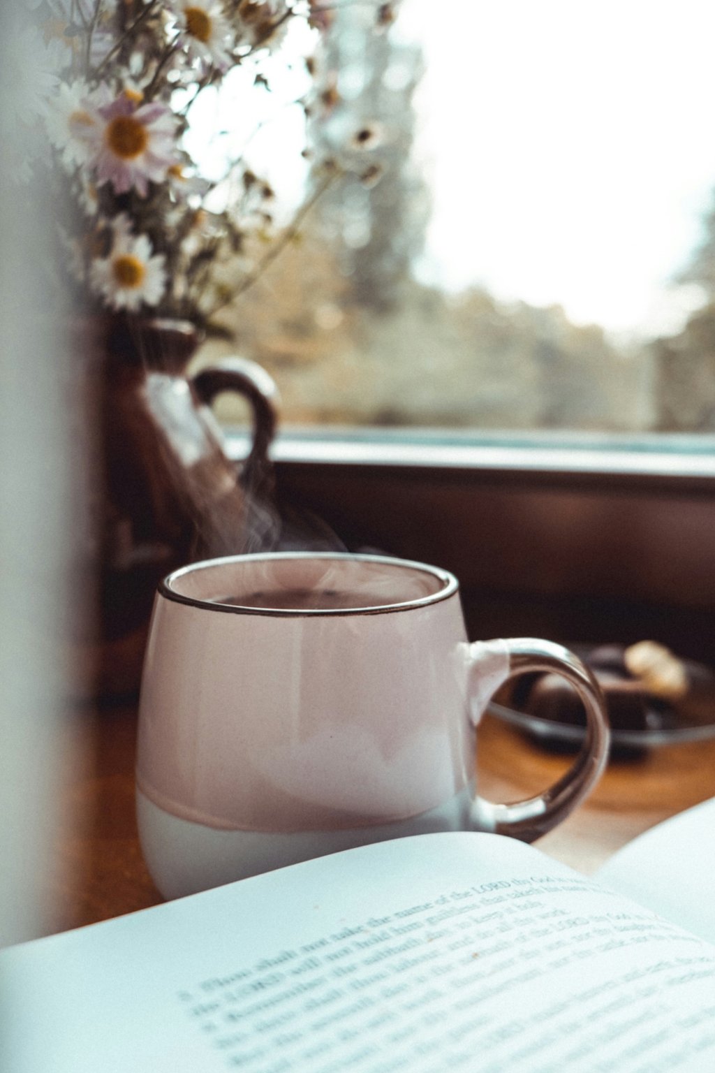A steaming cup of tea in a soft pink mug placed on a wooden table near an open book, with daisies in a vase in the background. A cozy, reflective scene for comfort.