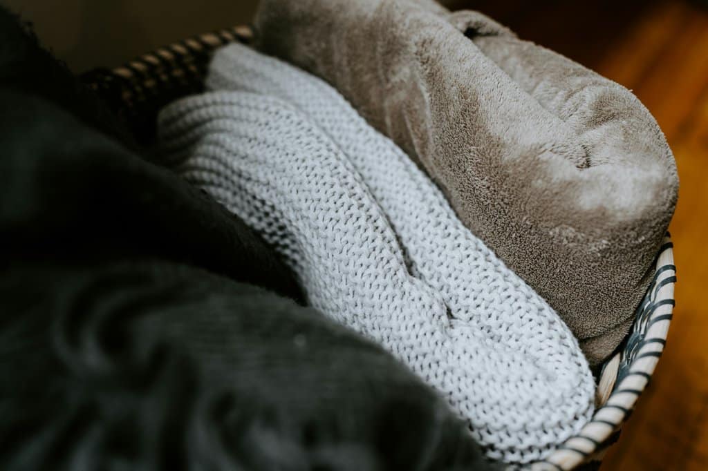 A basket filled with soft, folded blankets including a white knit throw and a beige plush blanket. Cozy textures that symbolize warmth and comfort during grief.